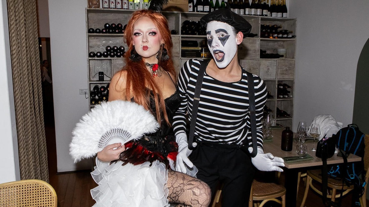 Two people in costumes, one with red hair and white makeup, the other in a striped outfit, in a room with shelves of bottles.